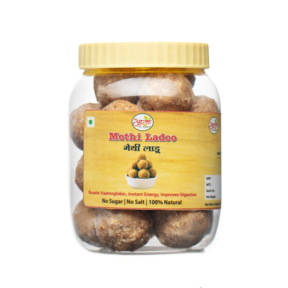 A clear plastic jar with a yellow lid contains round brown Methi Ladoo, a traditional Indian sweet made from fenugreek. The label highlights: No Sugar | No Salt | 100% Natural.